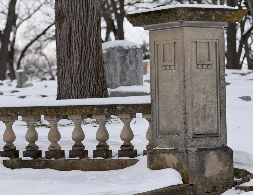 Stone railing and monument in a snowy cemetery, symbolizing winter dormancy and quiet renewal — photography by Ashley Lewis.